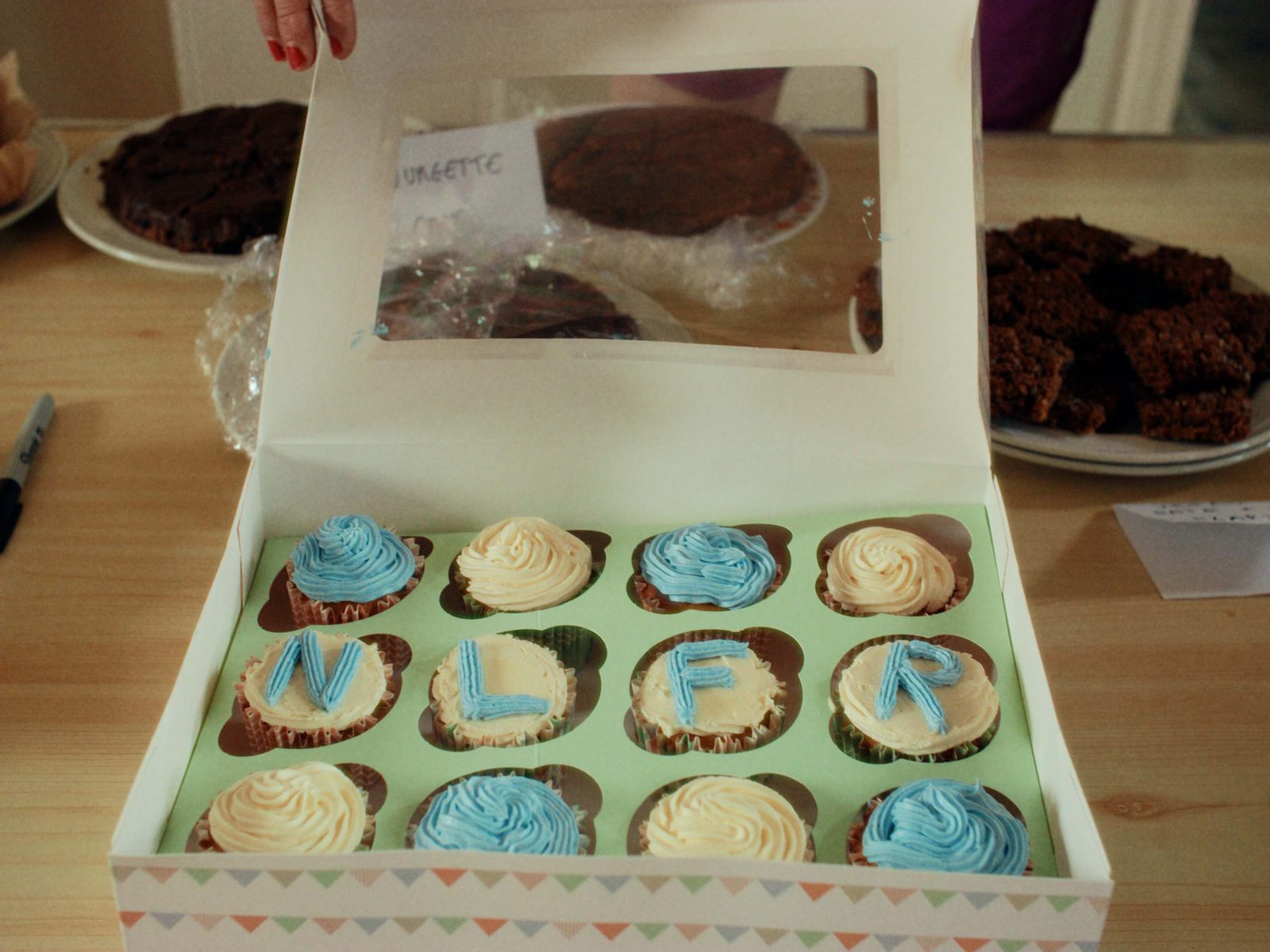 A box of homemade cupcakes iced with the letters NLFR beside prize cakes
